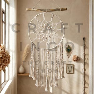 May include: A cream-colored macrame dream catcher with a circular design, suspended from a wooden branch. The dream catcher features intricate knotting and hanging tassels, creating a bohemian aesthetic. The text "THE CRAFT SHOP" is visible.