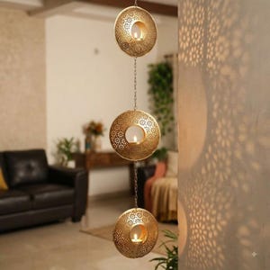 May include: A hanging gold-toned metal candle holder featuring three circular, perforated discs connected by thin chains. Each disc has a central circular opening designed to hold a small candle, creating intricate floral shadow patterns.