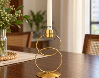 May include: A gold-toned metal candle holder featuring two interlocking circular rings mounted on a circular base. A single white taper candle is held in the top cup. The logo CRAFTINGO is visible on the upper ring of the candle holder.