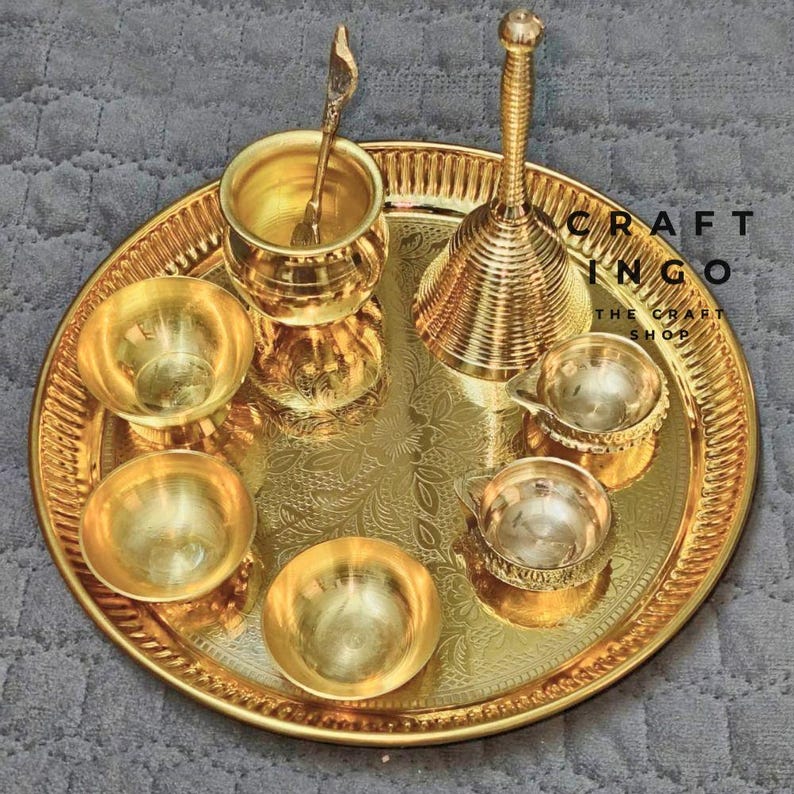 May include: A polished gold-tone brass set featuring a large circular tray with an engraved floral pattern, four small bowls, a tall bell with a ribbed handle, and a small cup containing a matching spoon, all arranged on a textured surface.