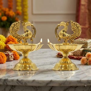 May include: A pair of polished gold-coloured metal oil lamps, each featuring a decorative peacock figure standing in the centre of a wide, scalloped bowl. The lamps are supported by a tiered, circular base and contain a small, lit wick.