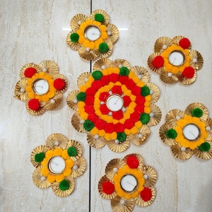 May include: A set of six decorative candle holders featuring gold pleated paper petals arranged in a flower shape. Each holder contains a white tea light candle and is accented with small red, yellow, and green pom-poms around the centre.