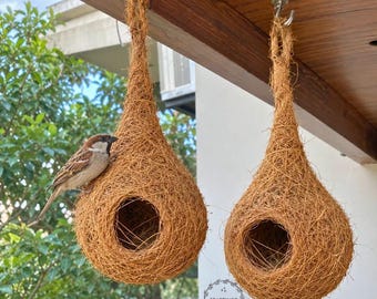 May include: Two teardrop-shaped bird nests made of brown woven coconut fiber hanging from a wooden beam. A small brown and white sparrow is perched on the edge of the left nest. The text CRAFTINGO THE CRAFT SHOP is visible below.