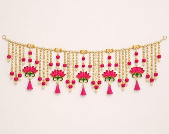 May include: A decorative wall hanging featuring a horizontal string of cream-colored beads with multiple vertical strands. These strands are adorned with small pink pom-poms, gold beads, and five prominent pink lotus flower charms with green bases.