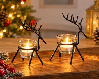 May include: Two black metal reindeer-shaped tea light candle holders, each featuring a clear glass cup holding a lit white tea light candle. The reindeer frames are constructed from thin metal rods with stylized antlers.