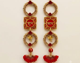 May include: Two vertical wall hangings featuring gold-toned circular wreaths adorned with small red flowers. Each piece includes a square red plaque with gold Hindi text, a gold Ganesha figure inside a wreath, and a red fabric tassel at the bottom.