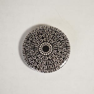 Puede incluir: Un botón redondo en blanco y negro con un patrón repetido de la palabra "ZUZU" en un diseño circular. El centro del botón presenta un patrón de círculos concéntricos más pequeños. El botón está sobre un fondo blanco.