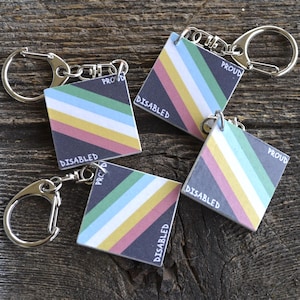 May include: Four square keychains featuring a diagonal striped pattern in green, light blue, white, gold, and pink. Each keychain has a black corner with the word PROUD printed in white and another black corner with the word DISABLED printed in white.