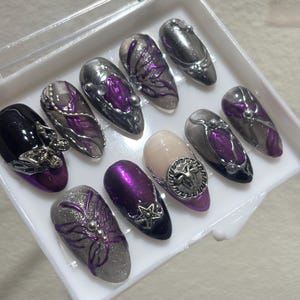 May include: A set of artificial nails in a clear plastic case. The nails feature a variety of designs, including silver glitter, purple accents, and metallic embellishments. Some nails have floral patterns, while others have geometric shapes.