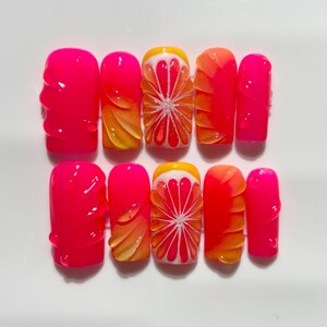Puede incluir: Un juego de diez uñas artificiales en un vibrante tono rosa neón. Algunas uñas presentan un diseño texturizado, mientras que otras muestran un degradado de amarillo y naranja. Dos uñas muestran un diseño detallado de pomelo con segmentos blancos y rojos.