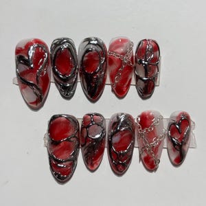 May include: A set of ten oval-shaped, press-on nails. Each nail features a red and white marbled design with silver metallic accents. Some nails have silver chain details, and others have a heart design.