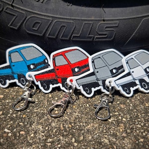 May include: Four miniature truck keychains in blue, red, gray, and white. Each keychain features a small truck design with black tires and a silver clasp. The keychains are arranged on a textured surface.