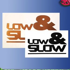 Puede incluir: Dos calcomanías rectangulares con las palabras "LOW & SLOW" en una fuente audaz y estilizada. Una calcomanía es de color cobre sobre un fondo crema, y la otra es negra sobre un fondo blanco. La imagen tiene un borde azul.