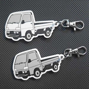 May include: Two keychains featuring a white and gray cartoon truck design. Each keychain has a silver-colored clasp. The trucks have a detailed outline and are set against a dark background.