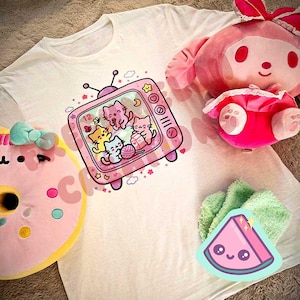 May include: A white t-shirt featuring a pink television graphic with cartoon cats. A pink plush toy, a pink donut plush, and a green watermelon sticker are also visible. The image has a soft, pastel aesthetic.