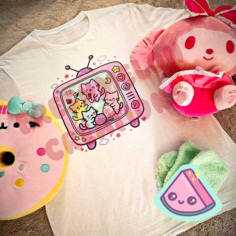 May include: A white t-shirt featuring a pink television graphic with cartoon cats. A pink plush toy, a pink donut plush, and a green watermelon sticker are also visible. The image has a soft, pastel aesthetic.