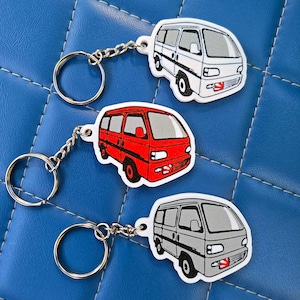May include: Three keychains featuring cartoon van designs in white, red, and gray. Each keychain has a silver chain and a round key ring. The vans have black tires and windows, with details like headlights and a front license plate.