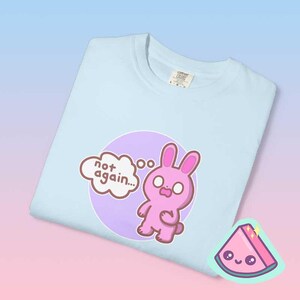 Close up folded pastel blue graphic tee showing pink bunny illustration and “not again” phrase, funny overwhelmed mood shirt