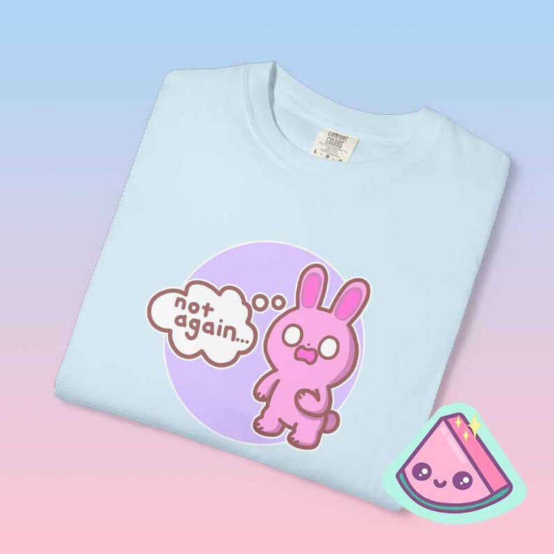 Close up folded pastel blue graphic tee showing pink bunny illustration and “not again” phrase, funny overwhelmed mood shirt