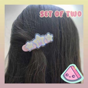 May include: A set of two star-shaped hair clips. The clips are light pink with three star designs in pastel yellow and blue. The text "SET OF TWO" is above the clips. A watermelon sticker is in the bottom right corner.