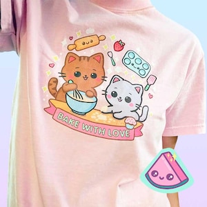 Puede incluir: Camiseta rosa claro con un dise&ntilde;o de dibujos animados de dos gatos horneando. Un gato mezcla en un cuenco azul, el otro sostiene una magdalena. El texto "Bake with Love" est&aacute; en una pancarta debajo de los gatos. Otros gr&aacute;ficos relacionados con la reposter&iacute;a tambi&eacute;n est&aacute;n presentes.
