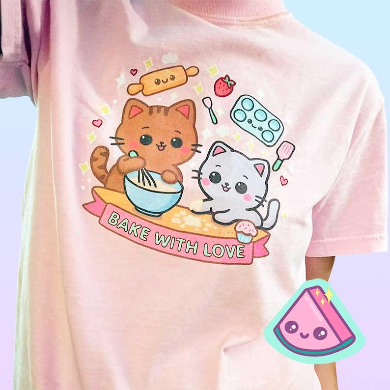 Puede incluir: Camiseta rosa claro con un dise&ntilde;o de dibujos animados de dos gatos horneando. Un gato mezcla en un cuenco azul, el otro sostiene una magdalena. El texto "Bake with Love" est&aacute; en una pancarta debajo de los gatos. Otros gr&aacute;ficos relacionados con la reposter&iacute;a tambi&eacute;n est&aacute;n presentes.