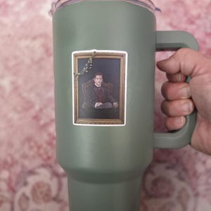 May include: A matte olive green insulated tumbler with a handle and a clear lid featuring a dark blue straw. A rectangular sticker is applied to the front, depicting a portrait of a person in a suit inside a gold frame with a small vine detail.