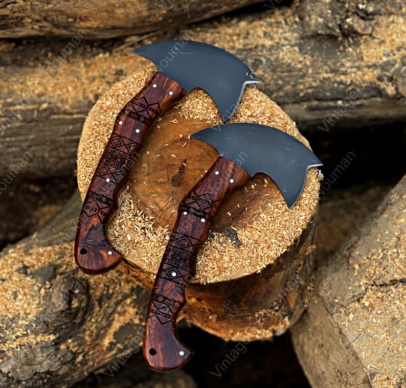 K&ouml;nnte beinhalten: Zwei Handbeile mit mattschwarzen, geschwungenen Klingen und dunkelbraunen Holzgriffen. Die Griffe weisen komplizierte, in das Holz eingravierte geometrische Muster auf, die durch kleine silberne Stifte entlang des Griffs akzentuiert sind.