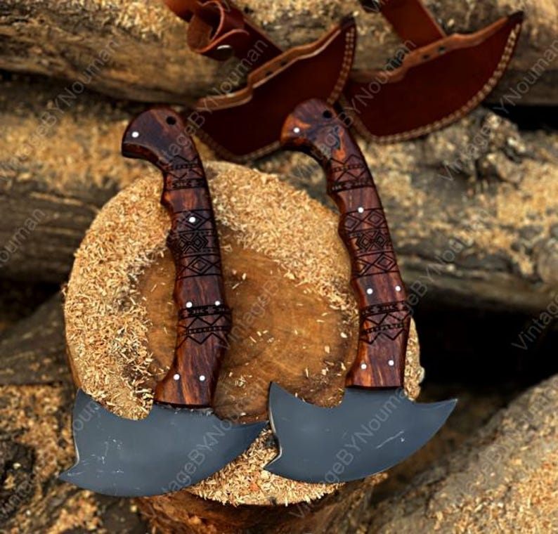 K&ouml;nnte beinhalten: Zwei Handbeile mit dunklen, matten Metallklingen und geschwungenen Holzgriffen, die mit komplizierten, lasergravierten geometrischen Mustern versehen sind. Jeder Griff ist mit kleinen Metallnieten befestigt. Zwei braune Lederscheiden liegen im Hintergrund.