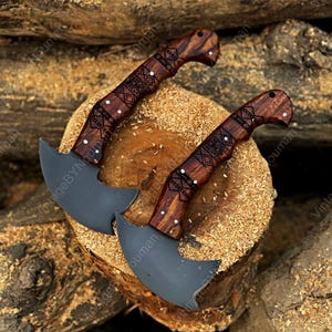 K&ouml;nnte beinhalten: Zwei passende Handbeile mit mattschwarzen, geschwungenen Klingen und dunkelbraunen Holzgriffen. Die Griffe sind mit komplexen geometrischen Mustern verziert und mit kleinen silbernen Stiften befestigt, was ein geschwungenes Griffdesign zeigt.