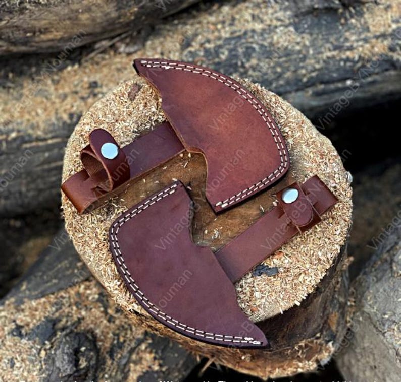K&ouml;nnte beinhalten: Zwei dunkelbraune Axtscheiden aus Leder mit wei&szlig;en N&auml;hten und silberfarbenen Metalldruckkn&ouml;pfen. Jede Scheide verf&uuml;gt &uuml;ber eine geschwungene Klingenschutzabdeckung und einen Riemen mit Druckknopfverschluss zur Sicherung des Axtkopfes.