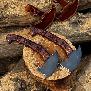 K&ouml;nnte beinhalten: Zwei halbmondf&ouml;rmige Ledermesser mit dunklen, matten Metallklingen und polierten Griffen aus dunklem Holz mit geometrischen Gravuren. Jedes Messer wird mit einer passenden braunen Lederscheide mit Riemen und Druckknopf geliefert.