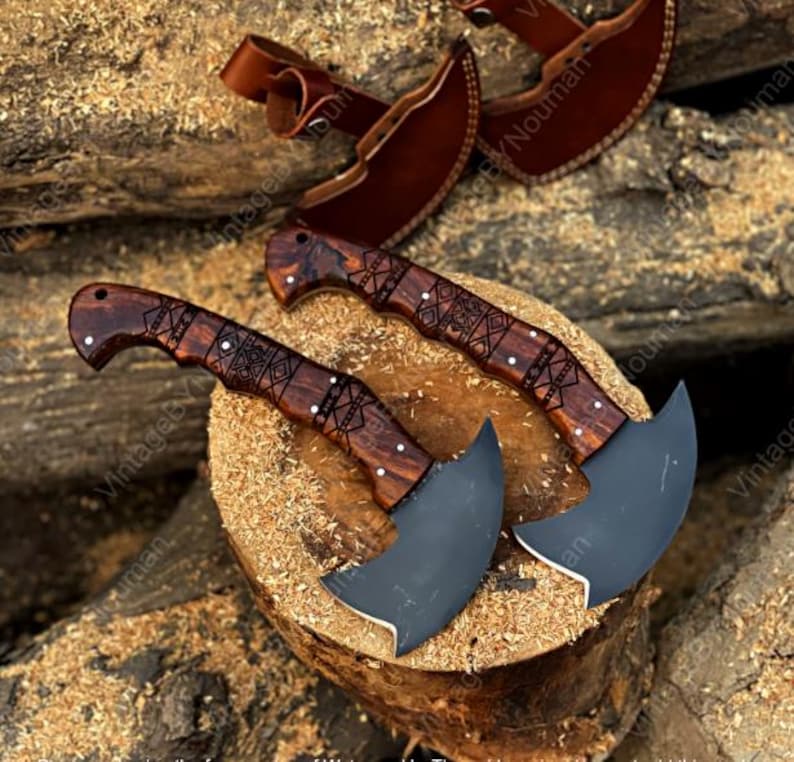 K&ouml;nnte beinhalten: Zwei halbmondf&ouml;rmige Ledermesser mit dunklen, matten Metallklingen und polierten Griffen aus dunklem Holz mit geometrischen Gravuren. Jedes Messer wird mit einer passenden braunen Lederscheide mit Riemen und Druckknopf geliefert.