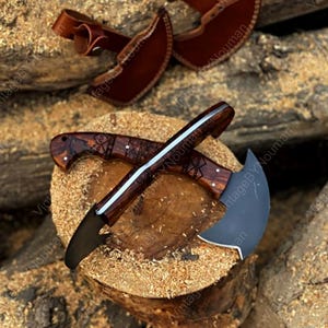 K&ouml;nnte beinhalten: Zwei halbmondf&ouml;rmige Messer mit dunklen Metallklingen und polierten Holzgriffen, die mit komplizierten geometrischen Gravuren versehen sind. Die Messer sind kreuzf&ouml;rmig angeordnet und werden von zwei braunen Lederscheiden mit Druckkn&ouml;pfen begleitet.