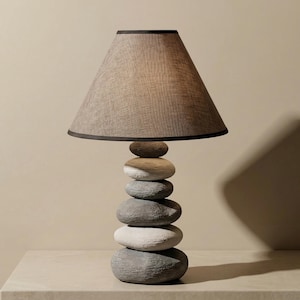Puede incluir: Lámpara de mesa decorativa con base de piedras apiladas y pantalla de tonos neutros. La base está compuesta por varias piedras grises y blancas, creando una estética equilibrada y natural. La pantalla es de color marrón claro con un borde marrón oscuro.