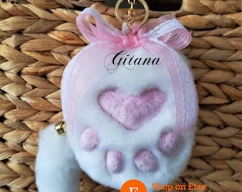 May include: A white and pink plush keychain with a heart and paw print design. The keychain has a gold-colored ring and a small bell. The text "Gitana" is written on the keychain. The Etsy shop name and handle are also visible.