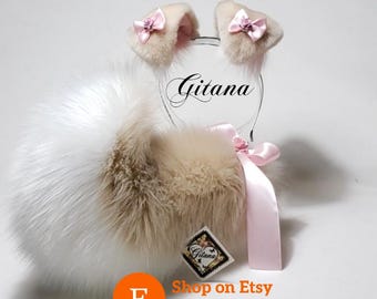 Peut inclure: Un serre-tête avec deux oreilles en fausse fourrure beige, chacune ornée d'un petit nœud en satin rose et d'une minuscule rose. Une queue en fausse fourrure blanche et beige est attachée avec un ruban en satin rose. Le texte Gitana est visible.