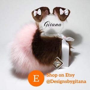 May include: A set of brown and light pink faux fur animal ears and a matching tail. The ears feature white satin bows with small pink centers. A white satin ribbon is attached to the tail. The text Gitana is visible on a clear circular stand.