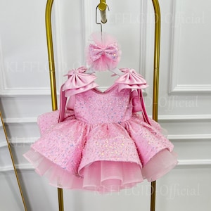 May include: A light pink dress featuring a sequined bodice with iridescent accents, large satin bows on the shoulders, and a voluminous layered tulle skirt. A matching pink hair accessory with a bow is attached to the top of the garment.