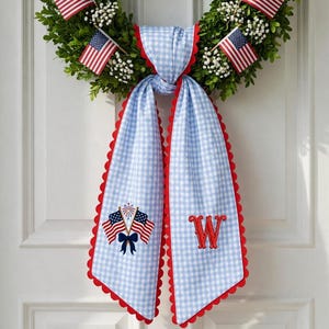 May include: A patriotic wreath with American flags and a blue and white gingham bow. The bow has red scalloped edges and embroidered details, including flags and the letter "W". The wreath is decorated with white flowers.