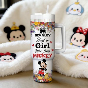 May include: A white Stanley travel mug with a handle, adorned with floral patterns and the text "Just a Girl Who Loves Mickey." It showcases a Mickey Mouse illustration and a straw. The mug is set against a backdrop of soft decorations.