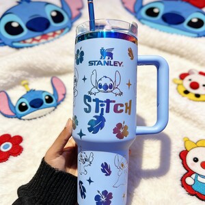 Peut inclure: Un gobelet Stanley bleu clair avec une poign&eacute;e, une paille et un couvercle transparent, orn&eacute; d'illustrations de Stitch et du mot "Stitch". Le design comprend des motifs floraux. Le gobelet est entour&eacute; de peluches sur le th&egrave;me de Stitch. Il mesure environ 18 cm de haut.