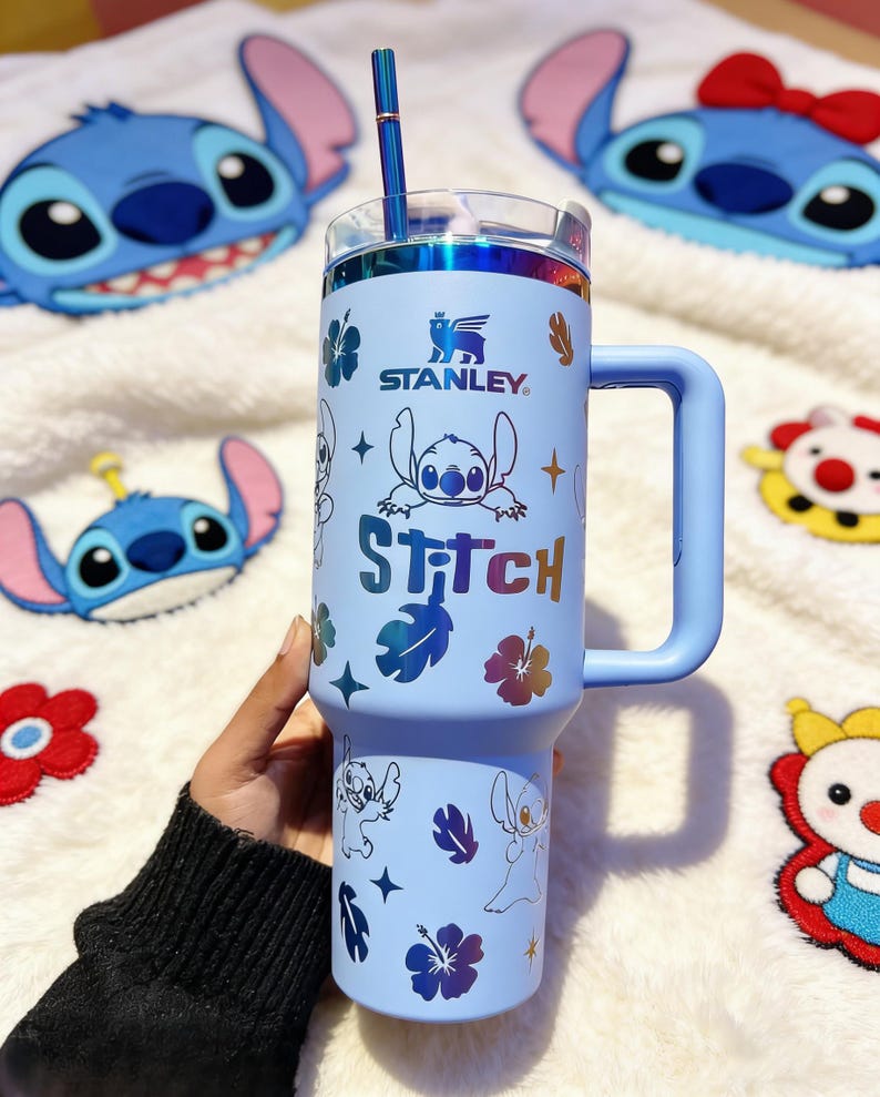 Peut inclure: Un gobelet Stanley bleu clair avec une poign&eacute;e, une paille et un couvercle transparent, orn&eacute; d'illustrations de Stitch et du mot "Stitch". Le design comprend des motifs floraux. Le gobelet est entour&eacute; de peluches sur le th&egrave;me de Stitch. Il mesure environ 18 cm de haut.