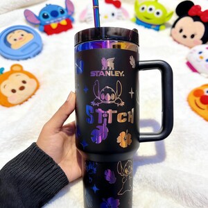 Peut inclure: Gobelet Stanley noir avec un motif Stitch et des accents iris&eacute;s. Le gobelet est dot&eacute; d'une poign&eacute;e, d'une paille et du mot "Stitch" imprim&eacute;. Le logo Stanley est &eacute;galement visible. Des jouets en peluche de personnages sont en arri&egrave;re-plan.