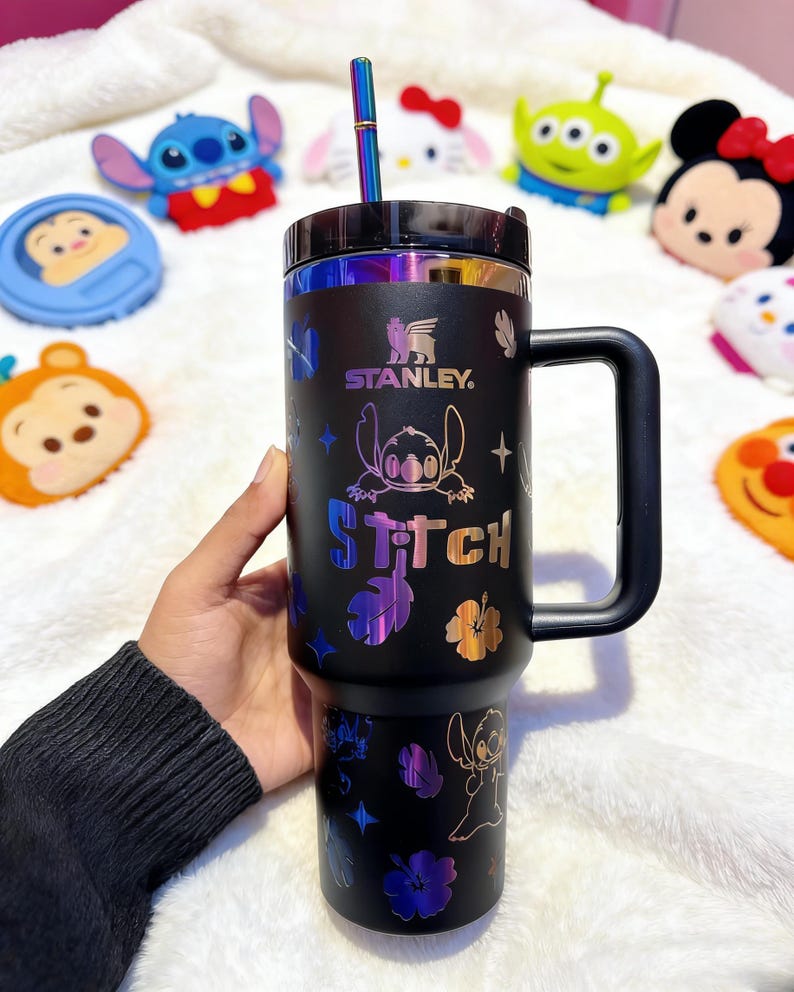 Peut inclure: Gobelet Stanley noir avec un motif Stitch et des accents iris&eacute;s. Le gobelet est dot&eacute; d'une poign&eacute;e, d'une paille et du mot "Stitch" imprim&eacute;. Le logo Stanley est &eacute;galement visible. Des jouets en peluche de personnages sont en arri&egrave;re-plan.