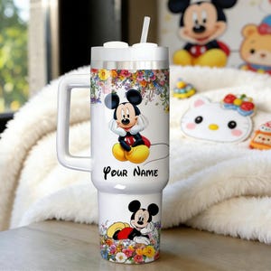 May include: White tumbler featuring Mickey Mouse with floral accents. The tumbler has a handle, a straw, and the text "Your Name". The design includes a cartoon Mickey Mouse image and colorful flowers.