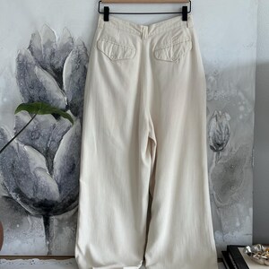 Puede incluir: Pantalones crema de pernera ancha con dos bolsillos traseros. Los pantalones cuelgan de una percha de madera frente a una pintura floral. La tela parece ser ligera.