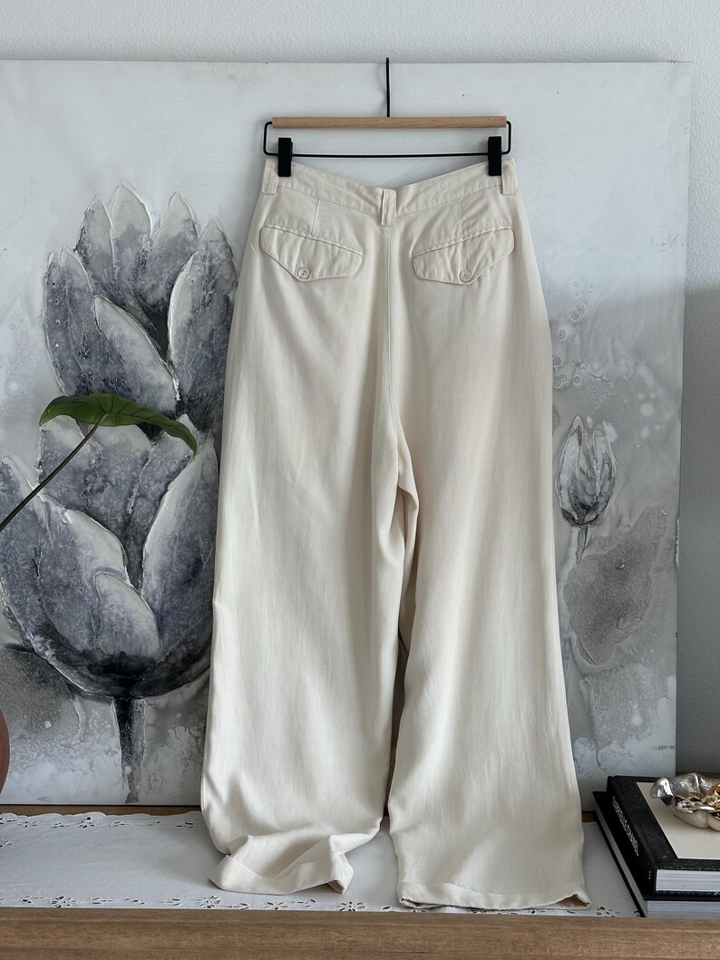 Puede incluir: Pantalones crema de pernera ancha con dos bolsillos traseros. Los pantalones cuelgan de una percha de madera frente a una pintura floral. La tela parece ser ligera.