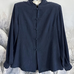Puede incluir: Blusa azul marino de manga larga con cuello mandarín y cierres de botón. La blusa presenta un cuello y puños con volantes. La tela tiene un sutil patrón texturizado. Una prenda de inspiración vintage.
