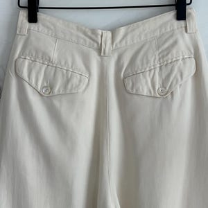 Puede incluir: Pantalones color crema con dos bolsillos traseros abotonados. Los pantalones est&aacute;n colgados de una percha negra con una barra de madera. La tela parece ser un material ligero, adecuado para climas c&aacute;lidos. Los pantalones tienen cintura alta y corte holgado.